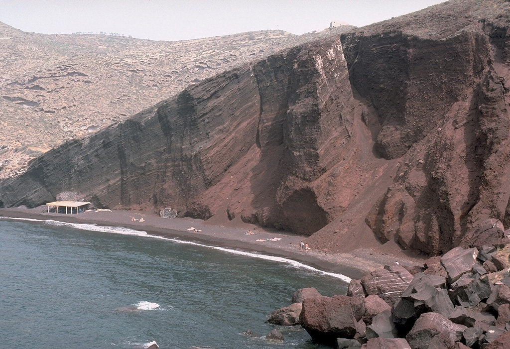 Wave erosion has exposed a cross section through bedded scoria units of the Cape Mavrorachidi scoria cone on the SW side of Thera Island. Behind the cone to the left are exposures of light-colored rhyodacitic tuffs of the Akrotiri Peninsula. A 451,000 +/- 27,000 BP (before present) Argon-Argon date was obtained from a lava flow from the Cape Mavrorachidi cone.  Photo by Lee Siebert, 1994 (Smithsonian Institution).