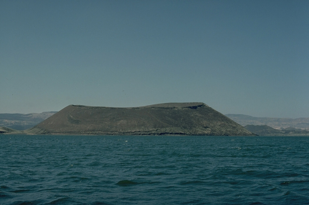 Naboiyoton tuff cone is seen here across Lake Turkana from the NE with The Barrier shield volcano in the background.  The steep slopes and wide crater of the tuff cone produce a flat-topped profile, which is typical of tuff cones.  This results from powerful phreatomagmatic eruptions produced by the interaction of magma and water.  The Barrier volcano draws its name from its prominent topographic position astride the East African Rift between Lake Turkana and the Sukota Trough to the south. Copyrighted photo by Katia and Maurice Krafft, 1978.