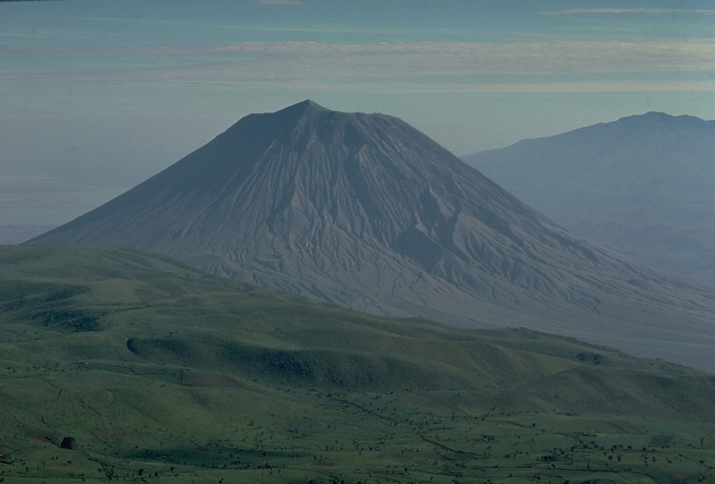 Symmetrical Ol Doinyo Lengai volcano is seen here from the slopes of Embagi volcano, to its SW.  The summit peak separates two craters, the northernmost (left-hand) of which has been historically active.  Ol Doinyo Lengai is renowned for its chemically distinctive products.  It is the only volcano known to have erupted carbonatitic tephras and lavas during historical time.  These sodium carbonate volcanic rocks are relatively poor in silica, the dominant chemical component of magmas erupted at nearly all of Earth's other volcanoes. Copyrighted photo by Katia and Maurice Krafft, 1976.