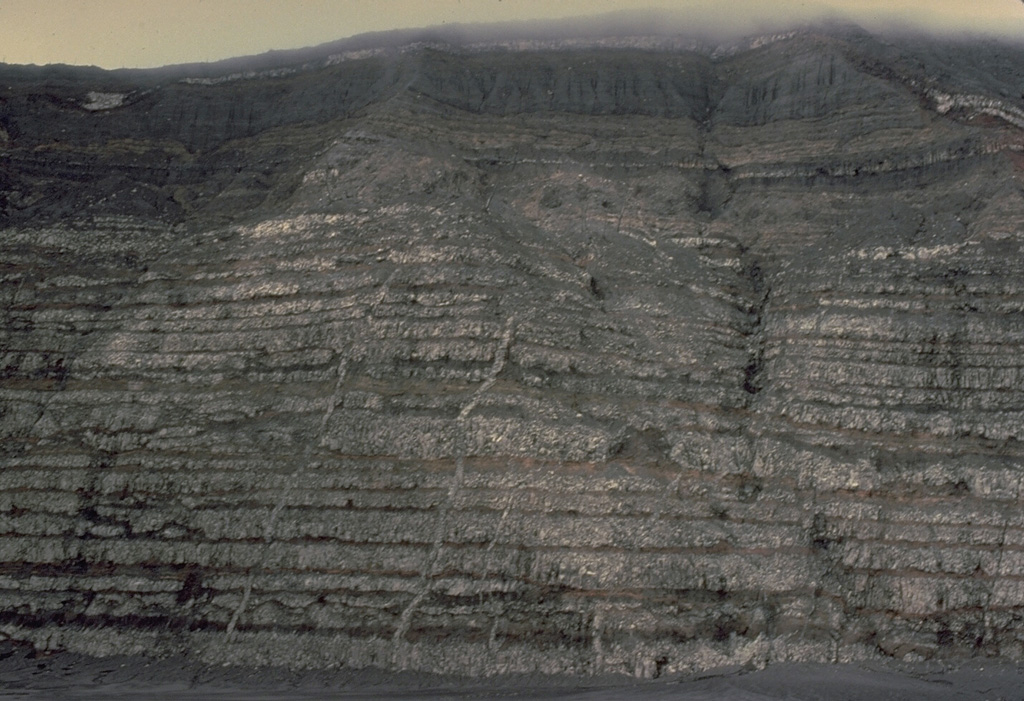 A strong explosive eruption at Karkar volcano in Papua New Guinea in 1979 stripped the southern caldera wall of vegetation, exposing a cross-section of the interior of the stratovolcano.  The 300-m-high caldera wall shows thin layers of light-colored lava flows separated by intervening pyroclastic layers, reflecting the repetitive effusive and explosive eruptions that built the volcano.  Narrow vertical dikes, the feeder conduits for lava flows, are visible at several locations in the caldera wall. Copyrighted photo by Katia and Maurice Krafft, 1979.