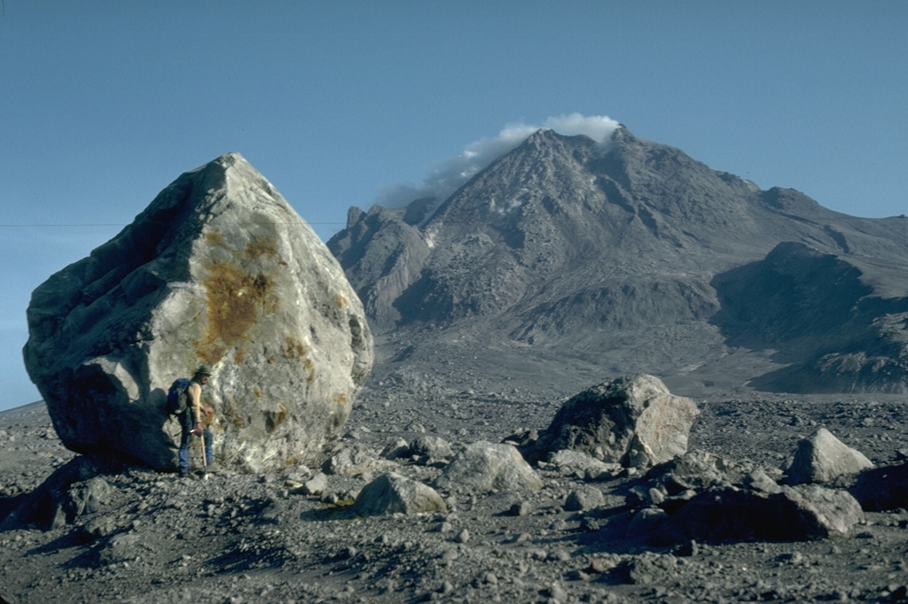 A volcanologist next to a 6-m-high block that was carried about 4 km down the north flank of Augustine volcano in Alaska during the 1976 eruption. Blocks of this size and larger are fragments of the summit lava dome that were carried within block-and-ash flows produced by periodic collapse of the growing dome. This photo was taken during a quiet phase of the 1986 eruption and shows the steaming summit lava dome. Photo by Harry Glicken, 1986 (U.S. Geological Survey).