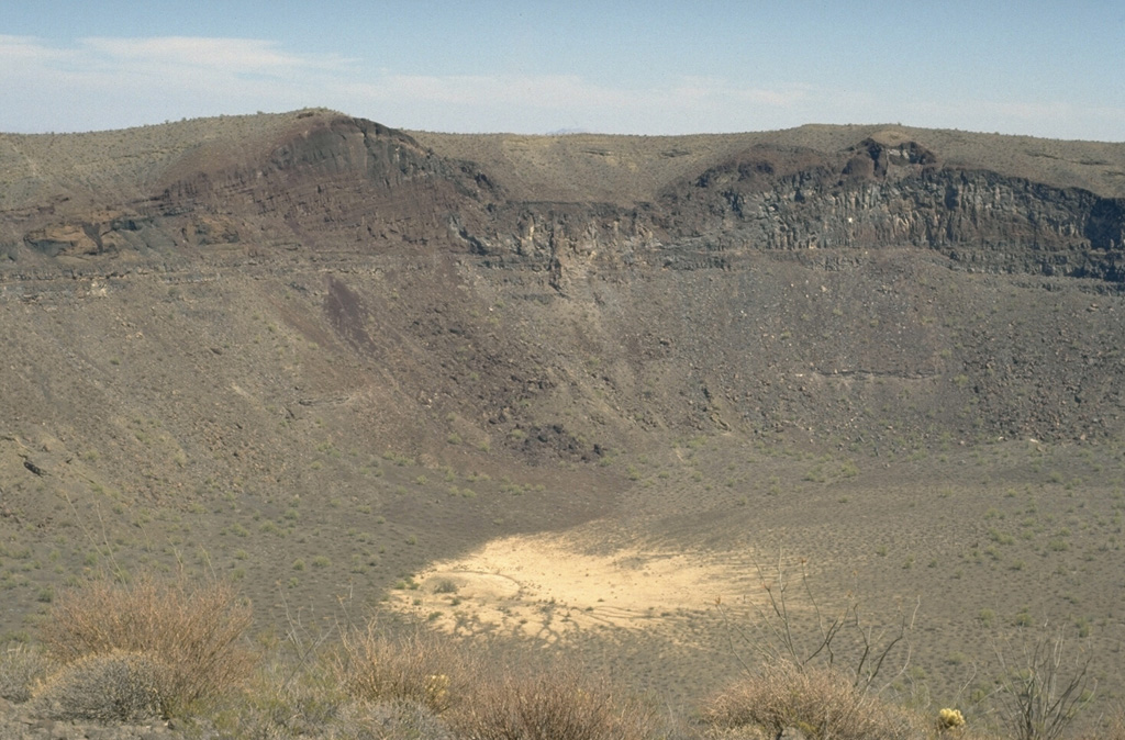 The SE wall of Cráter Elegante reveals a cross-section of a scoria cone that existed prior to explosive formation of the maar. The light-gray remnants of the sill intrusions are visible at the low point of the cone and below its right flank. The vent was located where the SE part of Cráter Elegante is now. The surface of the cone is mantled by pyroclastic surge deposits from the maar-forming eruption. Photo by Jim Luhr, 1996 (Smithsonian Institution).