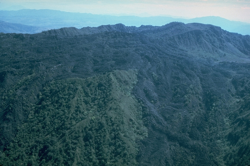 The El Norte lava flows, which cover much of the northern flank of Ceboruco, were erupted along buried ring fractures within nested calderas. The flows split into two lobes, a broad northern lobe that reached the base of the volcano and a narrower NW lobe (lower right). The southern wall of the inner caldera forms the irregular ridge behind the flows (below the distant haze), with the summit cone and lava flows visible at top right. Photo by Jim Luhr, 1980 (Smithsonian Institution).