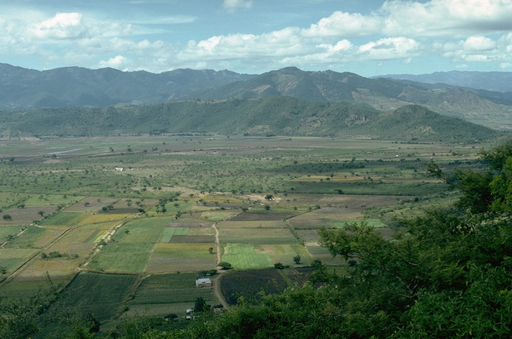 Retana caldera, between Suchitán and Tahual volcanoes, was once filled by Laguna Retana. The lake periodically became dry and was refilled in the 19th and 20th centuries and has now been drained to  access the rich soils on the lake floor. A canal drains the lake through a notch on its northern rim (far right). The caldera is seen here from its eastern rim, on the flank of Suchitán. Volcán Tahual is the forested volcano just behind the caldera wall and to the right. Photo by Lee Siebert, 1993 (Smithsonian Institution).