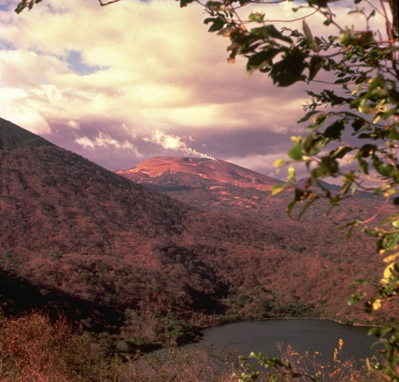 Las Pilas volcano (center), with a steam plume pouring from a vigorous fumarole on the SE side of the summit, is viewed here from the south rim of Laguna de Asososca, a 1.3 x 2 km wide maar on the southern end of Las Pilas volcanic complex.  The surface of Laguna de Asososca is less than 100 m above sea level.  At the left is the eastern flank of Cerro Asososca stratovolcano.   Photo by Jaime Incer, 1982.