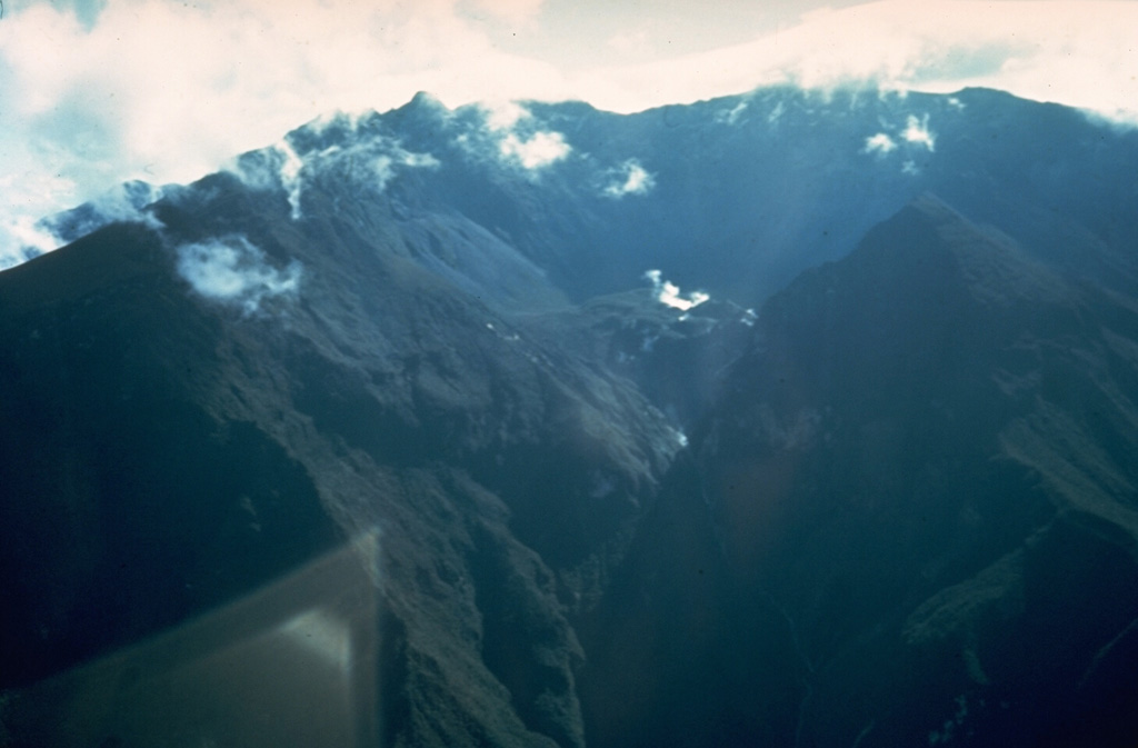 Fumaroles rise above a small crater (right center) at the base of the headwall of the 6-km-wide summit caldera of Guagua Pichincha volcano.  The breached caldera seen here from the east was formed about 50,000 years ago.  Subsequent late-Pleistocene and Holocene eruptions from the central vent in the breached caldera consisted of explosive activity with pyroclastic flows that accompanied periodic growth and destruction of a lava dome.   Photo by Minard Hall, 1975 (Escuela Politécnica Nacional, Quito).