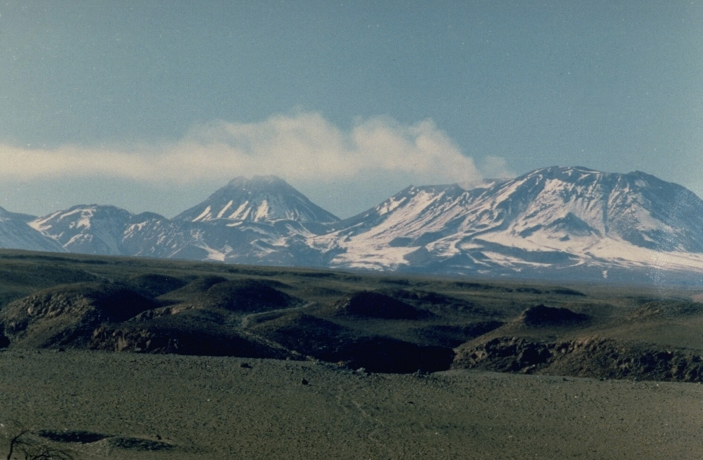 Volcán Láscar (right) is the most active volcano of the northern Chilean Andes.  A steam plume rises in 1986 from one of six overlapping summit craters capping the andesitic-to-dacitic stratovolcano, which is seen here from Toconao to the NW.   Volcán Aguas Calientes (left center), an older, higher stratovolcano 5 km to the east, displays a well-developed summit crater and a probable Holocene lava flow near its summit.   Frequent explosive eruptions have been recorded from Láscar since the mid-19th century. Photo by Paul King, MINSAL Corporation, 1986 (courtesy of Peter Francis, Open University).