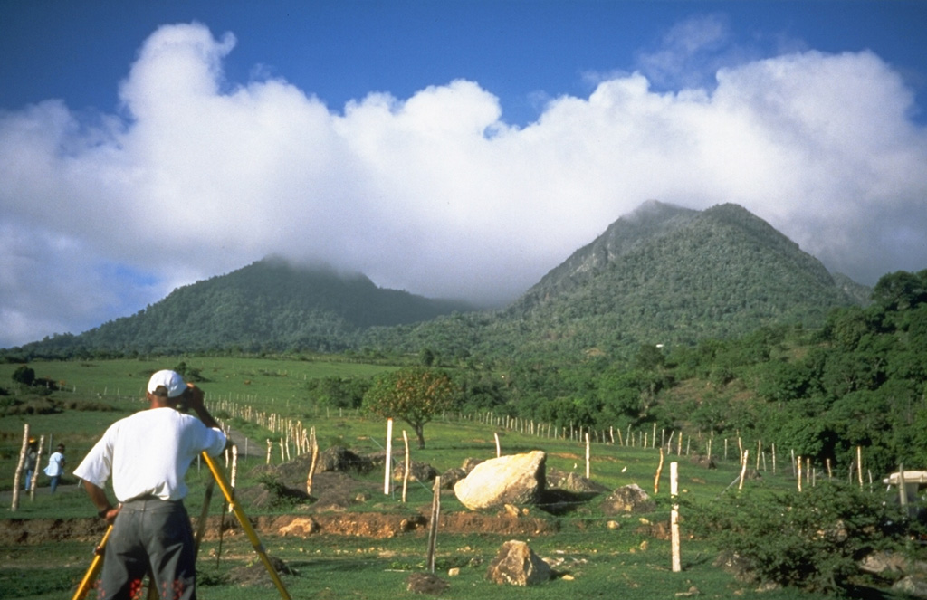 Volcanologist Richard Robertson of the Montserrat Volcano Observatory makes precision leveling measurements on the west flank of Soufrière Hills volcano on August 19, 1995.  Measurements such as these enabled scientists to track the progress of the eruption.  The boulders in the foreground are on the surface of pyroclastic-flow deposits from previous eruptions.  Gage's Mountain lava dome forms the peak at the right. Photo by Cynthia Gardner, 1995 (U.S. Geological Survey).