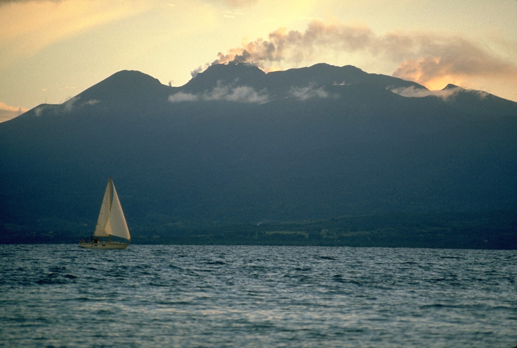 La Soufrière volcano, the highest in the West Indies, is a complex stratovolcano that dominates the southern end of Basse-Terre, the western half of the butterfly-shaped island of Guadeloupe.  A steam-and-ash column rises above the summit in this October 1976 view from the NE at Pointe-a-Pitre, on the isthmus connecting the island segments.  The peak at the left (above the sailboat), l'Echelle, is a young cinder cone; the summit itself is a 500-year-old lava dome, and remnants of older collapsed volcanoes are located to its right. Photo by Richard Fiske, 1976 (Smithsonian Institution).