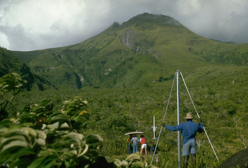 The summit of Pelée volcano rises immediately above scientists from the Mount Pelée volcano observatory and the Smithsonian Institution who are taking precision leveling measurements on the west flank.  This procedure, sometimes referred to as "dry tilt," detects deformation of the volcano that often precedes an eruption by measuring the precise differences in elevation between two stadia rods placed on fixed points.  This technique is part of monitoring efforts by the observatory to help detect future eruptions of this scenic, but deadly volcano. Photo by Lee Siebert, 1977 (Smithsonian Institution).