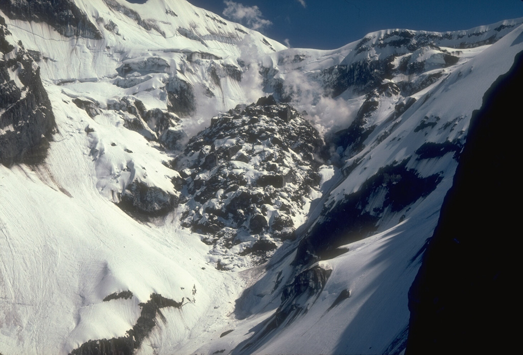 This view from the north shows the final lava dome of the 1989-90 eruption of Alaska's Redoubt volcano approximately one year after the end of the eruption. The dome measures 350-400 m across and contains around 10 million cubic meters of material. In this photo, snow is accumulating on the cooling lava blocks while hydrothermal activity continues to produce intermittent steam plumes. Periodic lava dome growth during the eruption was punctuated by strong explosions that destroyed earlier lava domes. Photo by Game McGimsey, 1991 (Alaska Volcano Observatory, U.S. Geological Survey).