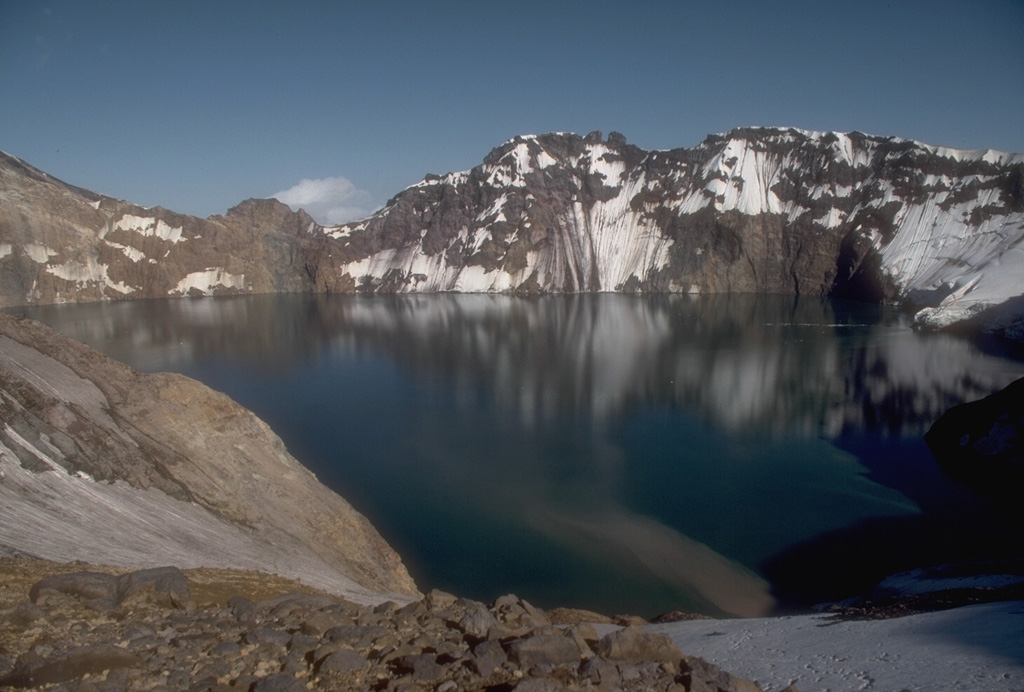 The 3 x 4.5 km Katmai caldera, seen here from its western rim, formed in 1912 by the hydraulic draining of magma away from Katmai to Novarupta 10 km W. Little if any eruptive activity took place at Katmai itself, which prior to collapse was a complex of four small overlapping stratovolcanoes. The 250-m-deep caldera lake covers a small lava dome and tuff ring that erupted on the caldera floor. Photo by Game McGimsey, 1990 (Alaska Volcano Observatory, U.S. Geological Survey).