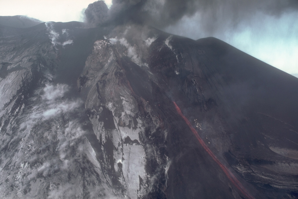 Incandescent lava flows down the side of a scoria cone in Veniaminof caldera on 7 October 1983 as ash plumes rise above the vent. This view from the SW shows the upper flanks of the cone, which is located on the west side of the glacier-covered caldera. Photo by Betsy Yount, 1983 (Alaska Volcano Observatory, U.S. Geological Survey).