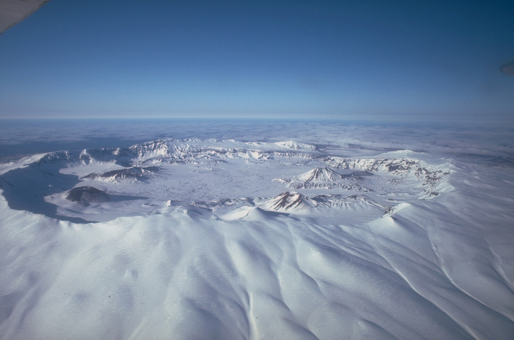 The summit of 35-km-wide Okmok has two largely overlapping 10-km-wide calderas. Both calderas formed by voluminous eruptions of tephra and pyroclastic flows during the Holocene, one about 8,250 years ago and the other about 2,400 years ago. Numerous cones and lava domes formed on the caldera floor and flanks. Historical explosive eruptions and lava flows have originated from cones within the caldera. Photo by John Reeder (Alaska Division of Geological & Geophysical Surveys).