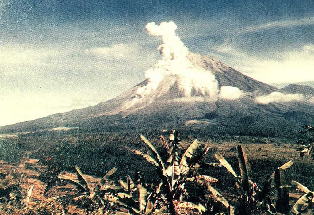 An eruption plume rises above the summit crater and a pyroclastic flow descends the southern flank of Semeru in this view from the Gunung Sanur Volcano Observation Post of the Volcanological Survey of Indonesia. The symmetrical stratovolcano is one of the most active in Java and has been in near-continual eruption since 1818. Frequent small-to-moderate explosive eruptions are occasionally punctuated by larger eruptions that produce pyroclastic flows and lahars that have reached the foot of the volcano. Photo courtesy of Volcanological Survey of Indonesia, 1992.