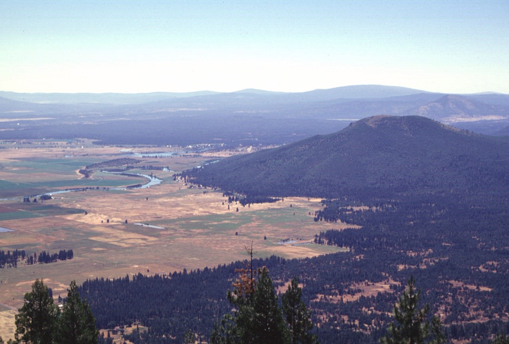 The broad forested area in the Falls River valley in the upper part of the photo beyond Saddle Mountain, the prominent forested peak at right-center, is the inconspicuous Big Cave shield volcano.  The low shield volcano fed lava flows that extend primarily to the north, towards the Pit River.  Big Cave is one of several poorly known young Quaternary volcanoes in northern California between Mount Lassen and Medicine Lake volcano.  This view looks to the SE from Soldier Mountain, with the town of Fall River Mills in the valley left of Saddle Mountain. Photo by Lee Siebert, 1998 (Smithsonian Institution).