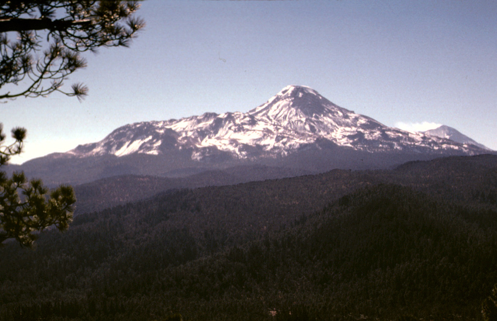 The profile of Iztaccíhuatl seen from the summit of Papayo volcano to the north. The northern base of the volcano is the Llano Grande edifice (the oldest edifice of Iztaccíhuatl) and the NW-flank La Trampa lava flows. The low-angle ridge extending to the NE (left) consists of the Teyotl dacite lava flows that erupted from a vent on the upper northern flank about 80,000 years ago. A gas plume can be seen at the summit of Popocatépetl to the far right. Photo by Lee Siebert, 1997 (Smithsonian Institution).