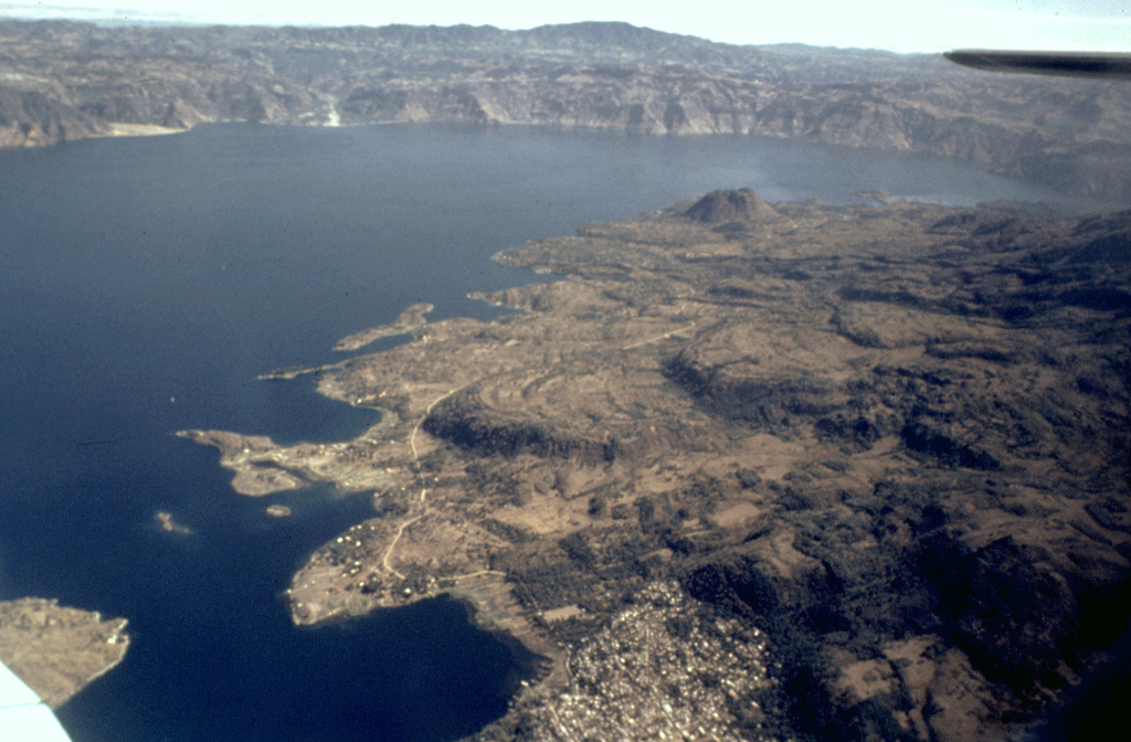 Lobate lava flows from Tolimán volcano form the irregular shore jutting into Lake Atitlán. The Cerro de Oro cone is on the near shore to the upper right, and the NE wall of Atitlán caldera rises about 1 km above the far side of the lake. The town of Santiago Atitlán (foreground) lies near the mouth of Santiago Bay. The buried margin of Atitlán I caldera, the first of three Miocene-Pleistocene calderas at Atitlán, lies approximately below Cerro de Oro; the boundary of Atitlán II caldera lies just below the bottom of the photo. Photo by Bill Rose, 1980 (Michigan Technological University).