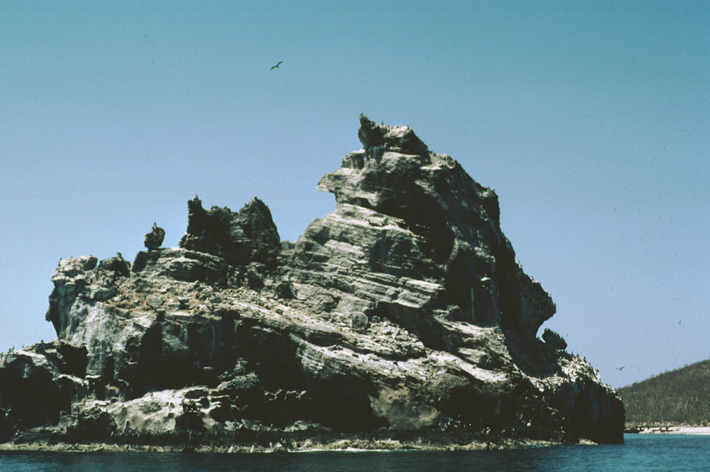 Wave erosion of a tuff cone off the western coast of Isla Isabel has produced the jagged profile of the 100-m-wide southernmost sea stack in the Islotes Las Monas. The three guano-covered cone remnants forming the Islotes Las Monas lie within about 200 m of the eastern coast of Isla Isabel, which can be seen to the right. Photo by Jim Luhr, 1999 (Smithsonian Institution).