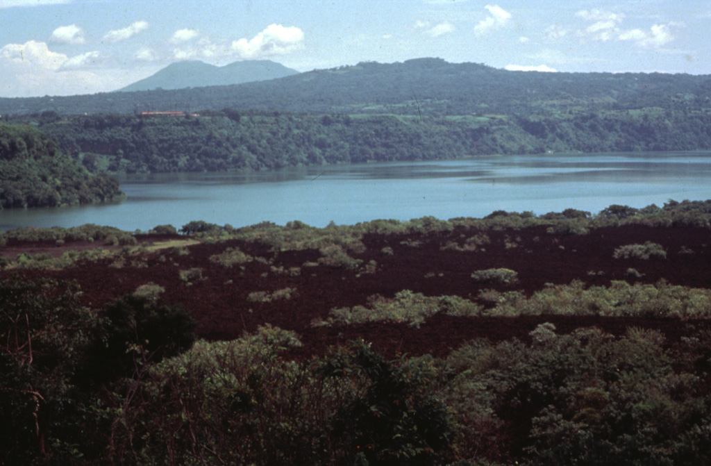 The sparsely vegetated lava flow in the foreground was emplaced during an eruption in 1772.  The flow originated from a vent on the north side of Old Masaya crater and traveled to the north.  One lobe passed through a notch in the northern caldera rim, while the lobe seen in this photo was deflected by the caldera rim and traveled to the SE into Lake Masaya, which is ponded against the SE caldera rim.  The twin-peaked stratovolcano in the distance is Mombacho volcano. Photo by Jaime Incer.