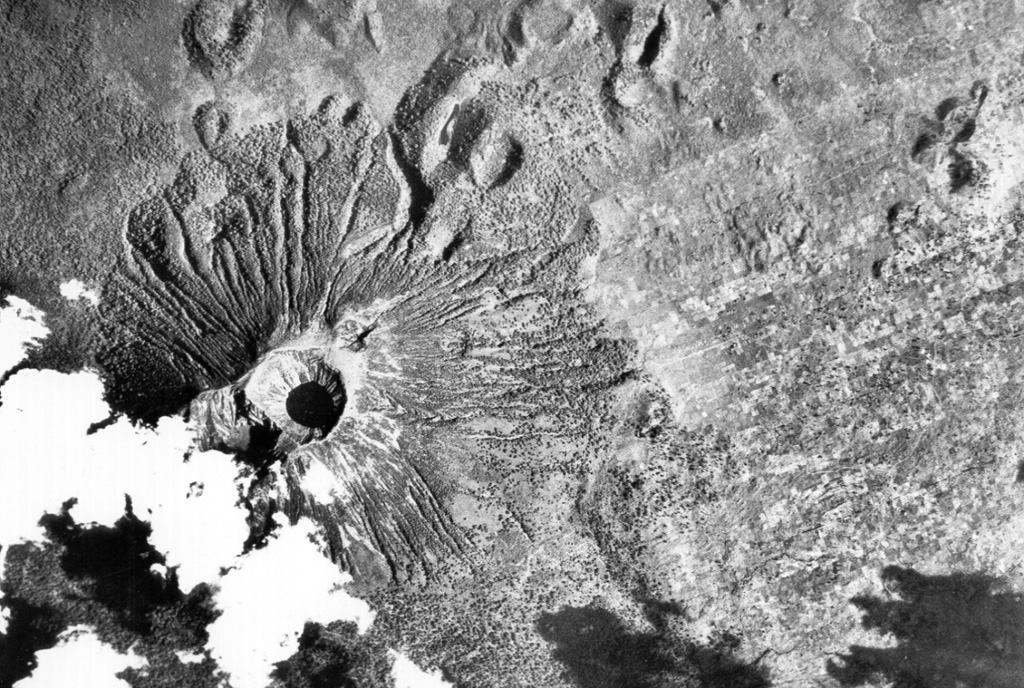 Visoke volcano is the NE-most of a cluster of large stratovolcanoes at the southern end of the Virunga Range. The volcano contains a 450-m-wide lake in its summit crater (left center). Lobate lava flows descend the flanks of the volcano. A NE-SW-trending fissure zone has produced many scoria cones NE of the volcano (top). An eruption in 1957 formed two small cones on the north flank, 11 km from the summit. Aerial photograph courtesy of Henry Luc Hody (Belgian ambassador).