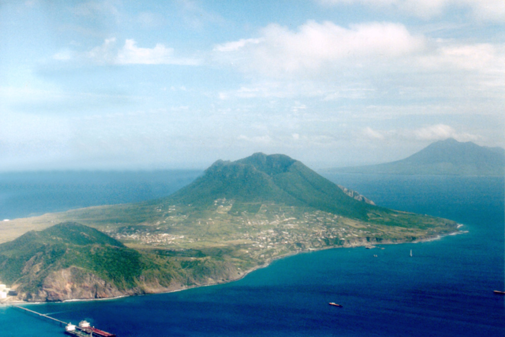 The Quill stratovolcano (center) forms the SE end of Statia (St. Eustatius) Island.  Pleistocene volcanic rocks form the hill at the lower left, and Mount Liamuiga volcano on St. Kitts (St. Christopher) Island rises across the strait at the right center.  A steep-sided 760-m-wide crater truncates the summit of The Quill, which was formed about 32,000-22,000 years ago by rhyolitic eruptions on a shallow-water limestone bank.  Pyroclastic-flow and -surge deposits, the last of which were erupted about 400 CE, blanket the slopes of the volcano.     Photo by John Shepherd, 2000 (Seismic Research Unit, University of West Indies).