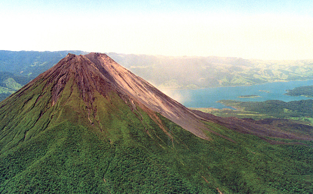 Below the vegetation on the NE Arenal flank is the A2 lava field. These voluminous flows cover the eastern flanks and are considered to have been erupted between about 1700 CE and the start of accurate historical records in Costa Rica in 1800 CE. The vegetation-free flank of the post-1968 cone form the right-hand side of the edifice, and recent lava flows from the 1968-2010 eruption are visible on the lower right-hand flanks with Lake Arenal in the background. Photo by Federico Chavarria Kopper, 1999.