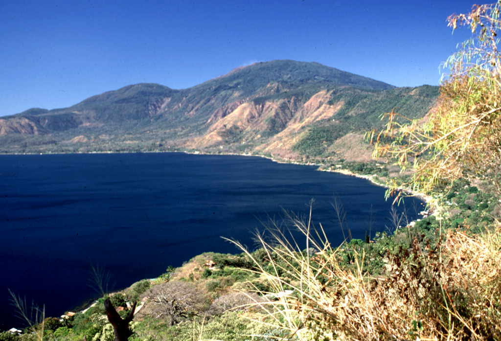 Coatepeque caldera is seen here from its northern rim, with Santa Ana volcano in the background. The caldera walls rise from 250 m to about 1 km above the lake and the stratigraphy is exposed in some areas. The rounded summit behind the caldera rim to the left is Cerro Verde, which was erupted along a NW-SE-trending fissure cutting through Santa Ana.  Photo by Lee Siebert, 2002 (Smithsonian Institution).