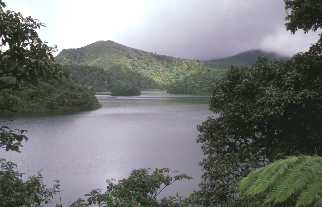 Freshwater Lake (L'Etang) lies in the moat between Micotrin lava dome and the eastern wall of the Wotten Waven caldera, partially visible in the background.  The 7 x 4.5 x wide caldera is elongated in an SW-NE direction, and it extends on the SW to near the capital city of Roseau.  The two coalesced lava domes of Micotrin straddle the NE rim of the caldera.  Strong geothermal activity persists in the caldera, the most prominent of which lies near the village of Wotten Waven along the River Blanc and contains numerous bubbling pools and fumaroles. Photo by Lee Siebert, 2002 (Smithsonian Institution).