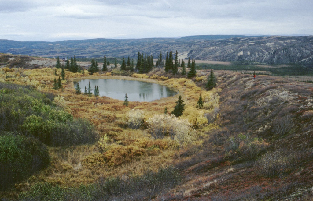 Moderate explosive eruptions radiocarbon dated to about 3,000 years ago formed two tuff rings in the central Alaska Range. A 300-m-wide ejecta blanket can be traced 1.6 km from the larger vent, seen in this photo. Ejecta on the crater rim consists of about 80% country rock fragments, with only 20% juvenile basaltic fragments. A geologist in a red jacket (right) and a helicopter (beyond the trees on the left side of the lake) on the crater rim, provide scale. Photo by Chris Nye (Alaska Division of Geological & Geophysical Surveys, Alaska Volcano Observatory).