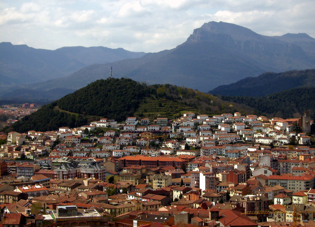 The forested Volcà Montolivet scoria cone rises above the city of Olot, as seen from the Volcà Montsacopa scoria cone to the NE. The cone is part of a large volcanic field named after the city and occupies the NE corner of Spain, south of the Pyrenees Mountains and about 90 km NNE of Barcelona. The latest dated eruption at Olot (also known as the Garrotxa volcanic field) occurred during the early Holocene and stratigraphic evidence suggests that more recent eruptions have occurred. Photo by Puigalder (Wikimedia Commons).