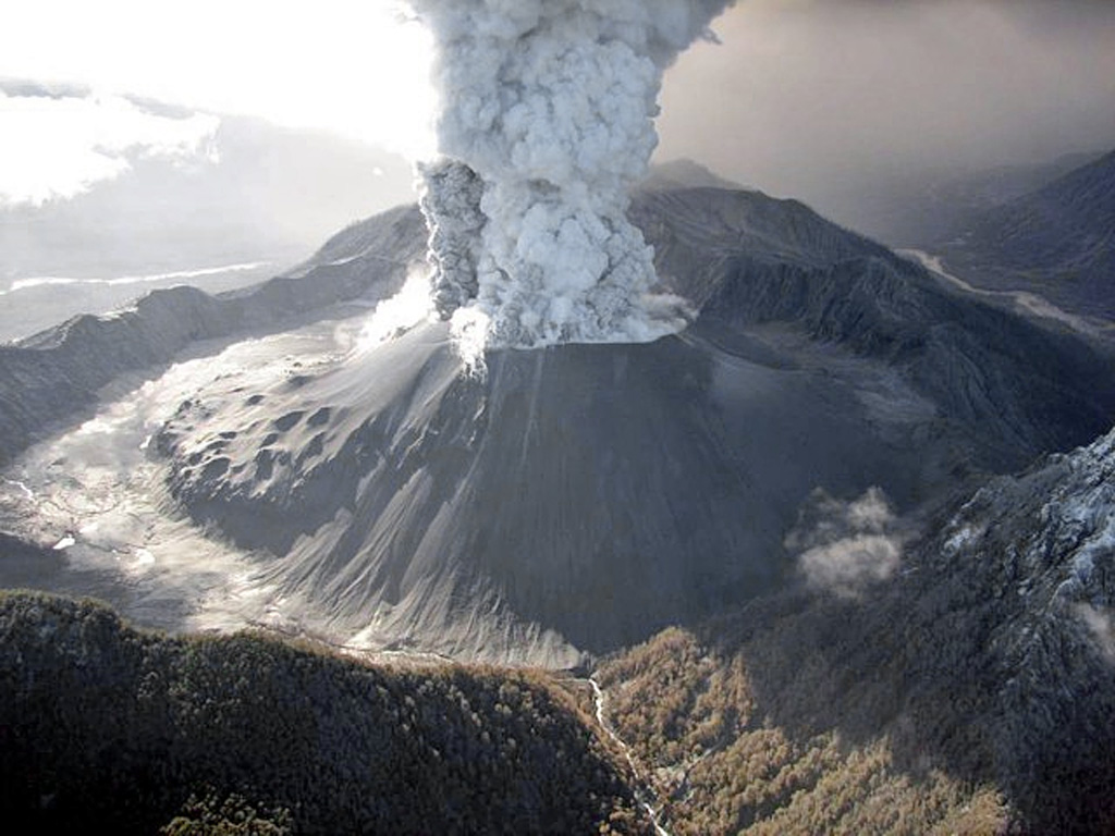 An eruption plume rises from a broad crater in a tephra cone atop the new and old dome complex in this May 26, 2008 helicopter view of Chaitén from the SW.  Lumpy areas on the middle to lower cone mark obsidian outcrops on the now buried older dome.  Burned vegetation is visible at the bottom center along the Blanco River.  A major explosive eruption at Chaitén volcano began on May 2, marking the first historical eruption of the volcano.  Mudflows destroyed much of the town of Chaitén.  Photo by Jeff Marso, 2008 (U.S. Geological Survey).