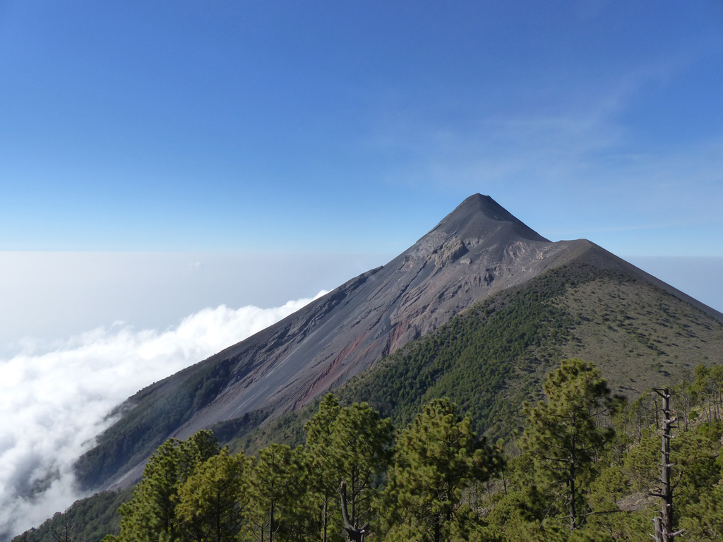 Fuego is seen here from Volcán Acatenango in February 2017. The ridge to the right of Fuego’s summit is a remnant of the older La Meseta edifice that underwent collapse approximately 9,000 years ago. The Barrancas Hondas and Las Lajas ravines that channel pyroclastic flows and lahars are visible on Fuego’s eastern flanks (left side); Honda is closer and deeper, with areas of red oxidation. Photo by Ailsa Naismith, 2017.